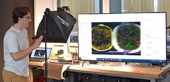 a person on the left views a large monitor at the right, which displays two pots of vegetation that have been scanned and analyzed by SUSHI